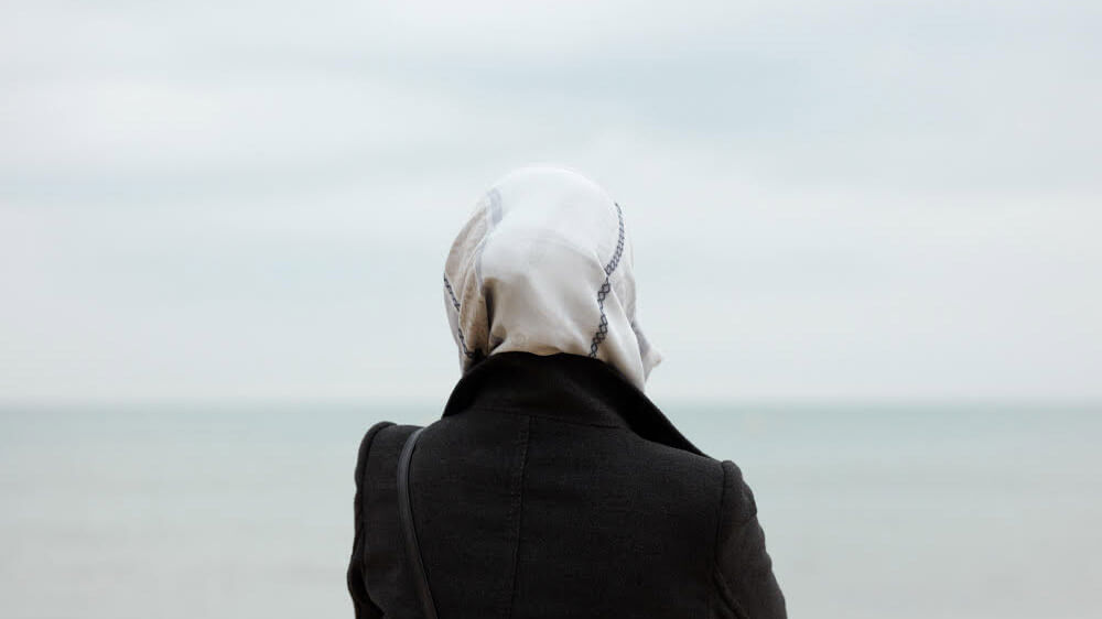 Woman in black coat and white head scarf stands with back to camera looking towards the sea
