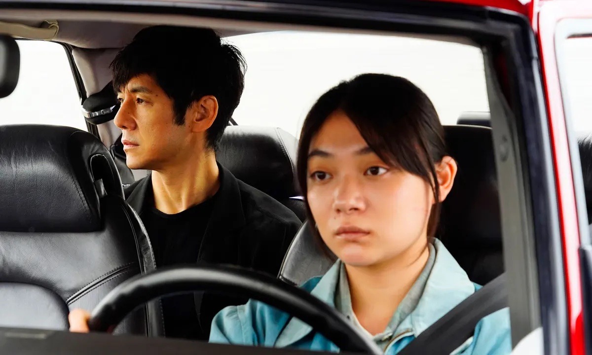 A Japanese woman is sat in the drivers seat in a red car holding the steering wheel. There is a man in the back on the right hand side looking out the window.