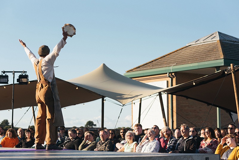 Actor Omar Bynon on stage, who played Decius / Soothsayer in Shakespeare’s Globe on Tour performance of Julius Caesar. He has his hands on the air looking at the audience with a tambourine in one hand.