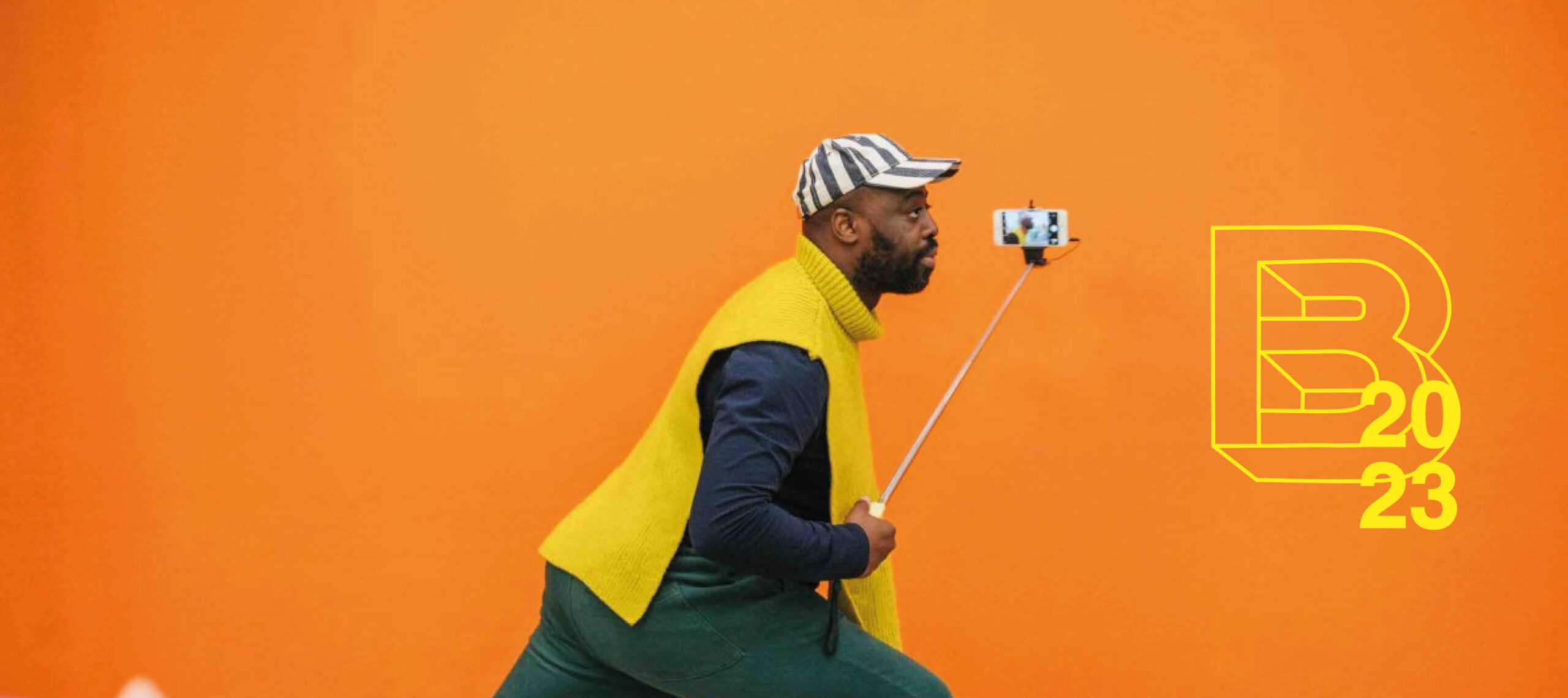 A side profile photograph of the artist Harold Offeh holding a selfie stick whilst lunging. There is a bright orange background, he is wearing a yellow sleeveless jumper, green trousers and a striped grey and white cap. In the right corner of this banner image is a yellow png of the BEAF 23 logo.